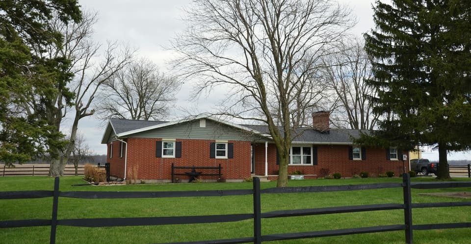 A single-storey red-brick ranch house behind a black rail fence, with bare trees and a lawn